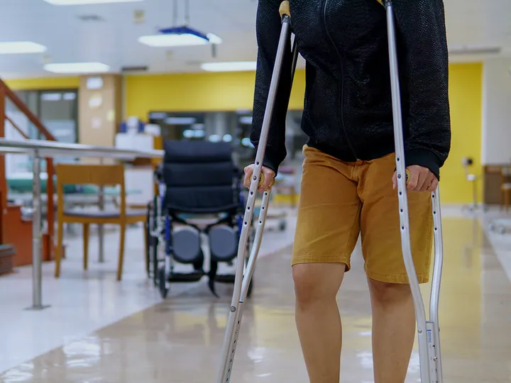 Pessoa caminhando de muletas no corredor de uma clínica de reabilitação, com cadeira de rodas ao fundo