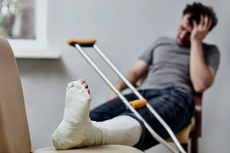 Homem sentado com a perna engessada apoiada e muletas ao lado, com a mão na cabeça, preocupado